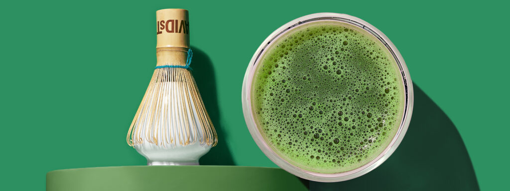 A bird's-eye view of frothy green matcha next to a bamboo whisk on a whisk holder against a dark green background.
